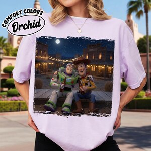 May include: A light purple t-shirt featuring a graphic print of Buzz Lightyear and Woody sitting together on a wooden platform at night in a western town setting. The text Comfort Colors Orchid is printed in a black oval in the upper left corner.