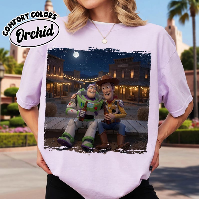 May include: A light purple t-shirt featuring a graphic print of Buzz Lightyear and Woody sitting together on a wooden platform at night in a western town setting. The text Comfort Colors Orchid is printed in a black oval in the upper left corner.