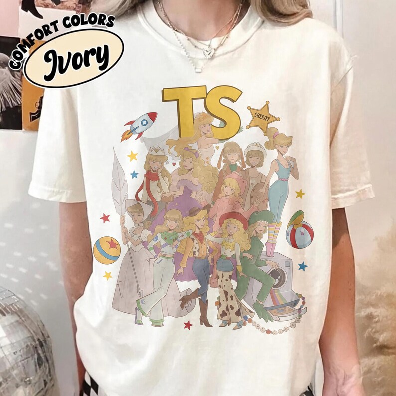 May include: An ivory short-sleeve t-shirt featuring a graphic print of various animated characters dressed in costumes, the letters TS in yellow, a sheriff star, a rocket, and colorful balls. The shirt has a crew neckline and a relaxed fit.