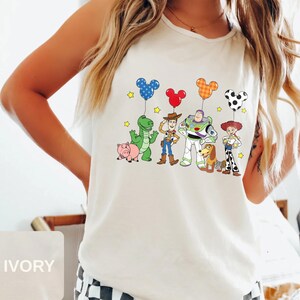 May include: Ivory tank top featuring a colorful graphic of Toy Story characters holding Mickey Mouse-shaped balloons. The design includes a green dinosaur, a pink pig, Woody, Buzz Lightyear, Jessie, and a dog. Yellow stars are scattered around the characters.