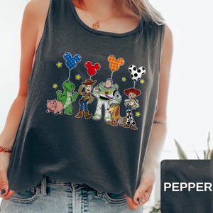 May include: Dark gray tank top featuring a colorful graphic of Toy Story characters holding Mickey Mouse-shaped balloons. The characters include Woody, Buzz Lightyear, and Jessie. The word "PEPPER" is printed on a black rectangle.