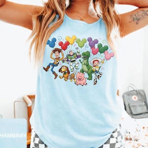 May include: A light blue sleeveless tank top featuring a colorful graphic print of Toy Story characters including Woody, Buzz Lightyear, Rex, Slinky Dog, Hamm, and Jessie. The characters are surrounded by a cluster of multi-colored Mickey Mouse balloons.