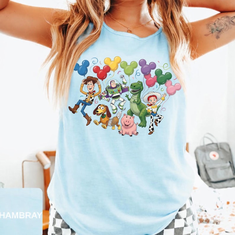 May include: A light blue sleeveless tank top featuring a colorful graphic print of Toy Story characters including Woody, Buzz Lightyear, Rex, Slinky Dog, Hamm, and Jessie. The characters are surrounded by a cluster of multi-colored Mickey Mouse balloons.