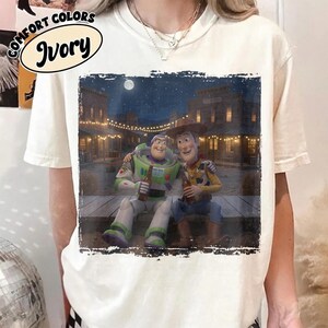 May include: An ivory-colored short-sleeve t-shirt featuring a graphic print of Buzz Lightyear and Woody from Toy Story sitting on a wooden dock at night. The scene includes a western-style town in the background under a full moon and string lights.