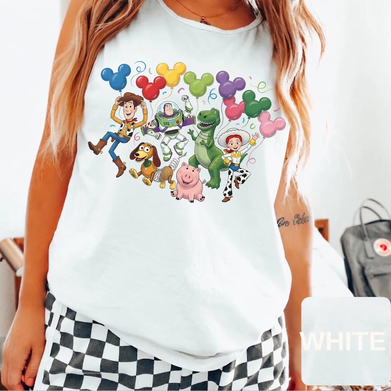 May include: A white sleeveless tank top featuring a colorful graphic print of various animated characters including a cowboy, a space ranger, a green dinosaur, a pig, a cowgirl, and a dog, all holding multi-colored balloons shaped like mouse ears.