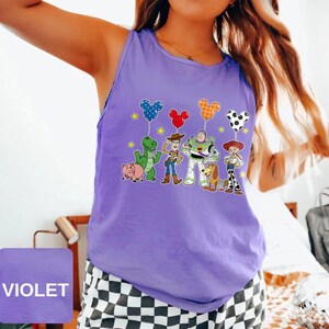 May include: A violet tank top featuring a graphic of Toy Story characters and Mickey Mouse-shaped balloons. The design includes Woody, Buzz Lightyear, and other characters from the animated film. The word "VIOLET" is printed on a square in the lower left corner.