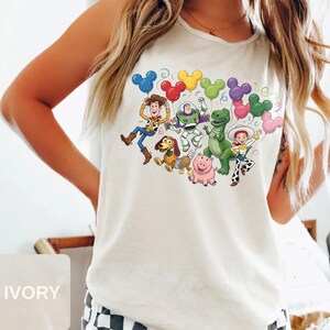 May include: An ivory sleeveless tank top featuring a colorful graphic print of various animated characters including a cowboy, a space ranger, a dinosaur, a pig, a dog, and a cowgirl, all holding multi-colored Mickey Mouse-shaped balloons.