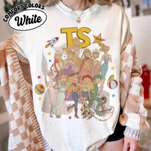 May include: A white crew neck t-shirt featuring a large graphic print of various animated characters in diverse outfits, including a space suit, cowboy attire, and gowns. The text TS appears in large yellow letters above the characters.