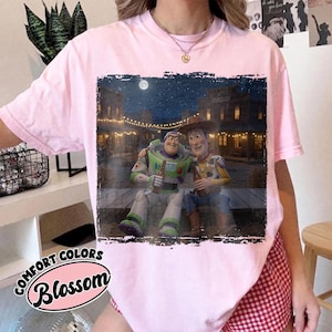 May include: A light pink short-sleeve t-shirt featuring a rectangular graphic print of Buzz Lightyear and Woody sitting together on a wooden porch at night in front of a western-style town with string lights and a full moon in the dark sky.