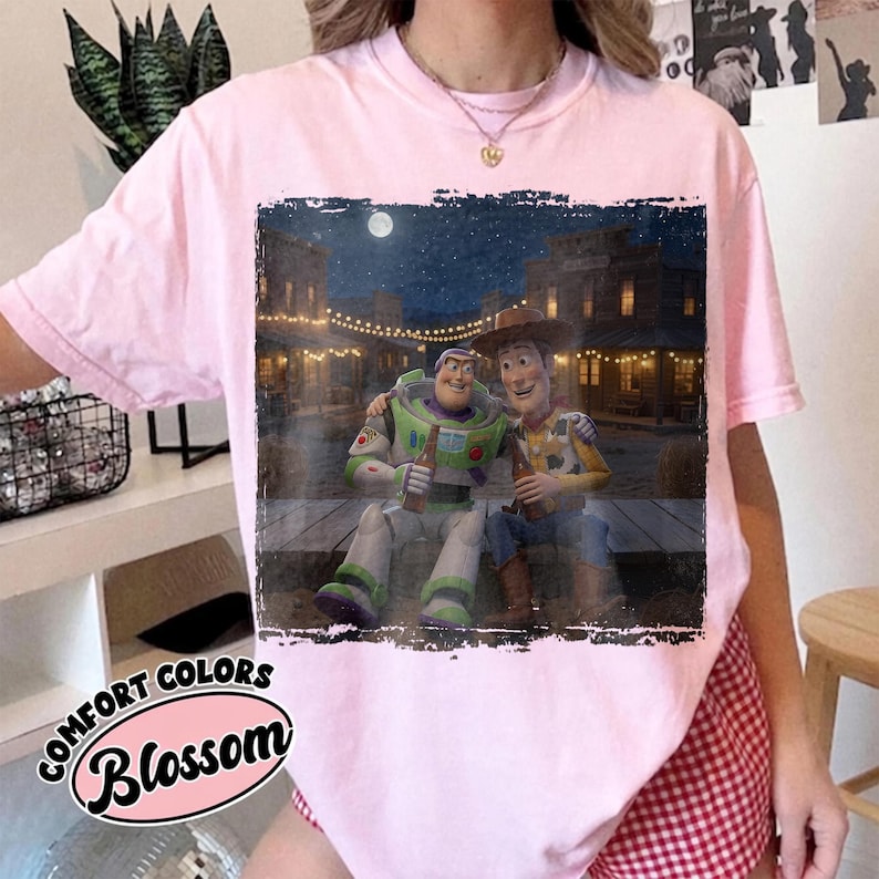 May include: A light pink short-sleeve t-shirt featuring a rectangular graphic print of Buzz Lightyear and Woody sitting together on a wooden porch at night in front of a western-style town with string lights and a full moon in the dark sky.