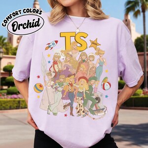 May include: A light purple short-sleeve t-shirt featuring a graphic print of various animated characters in diverse outfits, including a rocket, a sheriff badge, and balls. The text "COMFORT COLORS", "Orchid", and "TS" are printed on the shirt.