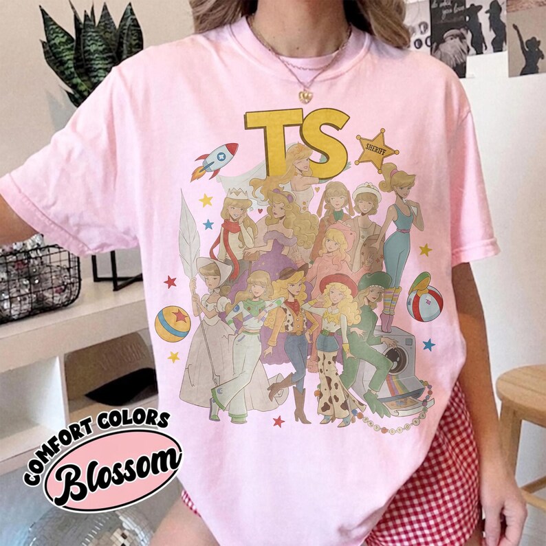 May include: A light pink short-sleeve t-shirt featuring a large graphic print of various animated characters in diverse outfits. The text TS appears in large yellow letters at the top, accompanied by a sheriff star, a rocket, and colorful stars.