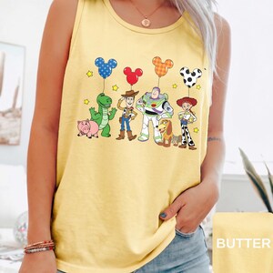 May include: Yellow tank top featuring a cartoon graphic of Toy Story characters. The characters are holding Mickey Mouse-shaped balloons. The word "BUTTER" is in a yellow rectangle in the bottom right corner.