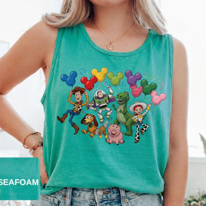 May include: A seafoam green sleeveless tank top featuring a graphic print of Toy Story characters including Woody, Buzz Lightyear, Rex, Jessie, Slinky Dog, and Hamm. The characters are surrounded by colorful Mickey Mouse-shaped balloons.