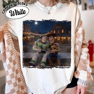 May include: A white t-shirt featuring a graphic print of Buzz Lightyear and Woody sitting together on a wooden porch at night. The scene includes a western-style town background with string lights and a full moon. The text Comfort Colors White is visible.