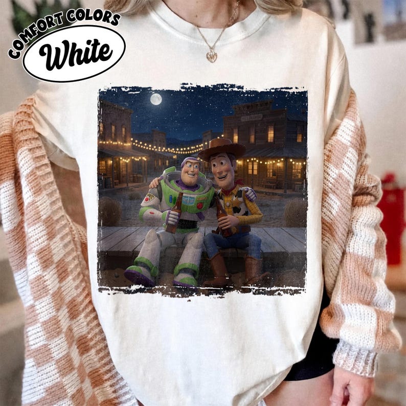 May include: A white t-shirt featuring a graphic print of Buzz Lightyear and Woody sitting together on a wooden porch at night. The scene includes a western-style town background with string lights and a full moon. The text Comfort Colors White is visible.