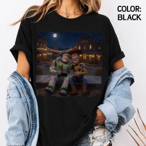 May include: A black t-shirt featuring a graphic print of Buzz Lightyear and Woody sitting together on a wooden porch in a western town at night under a full moon. The text COLOR: BLACK is printed in the upper right corner of the image.