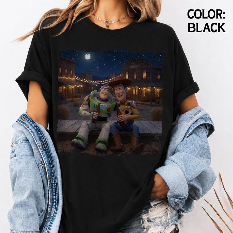 May include: A black t-shirt featuring a graphic print of Buzz Lightyear and Woody sitting together on a wooden porch in a western town at night under a full moon. The text COLOR: BLACK is printed in the upper right corner of the image.
