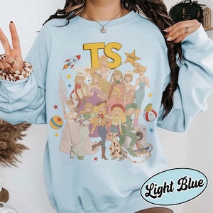 May include: A light blue crewneck sweatshirt featuring a large graphic print of multiple animated characters in various outfits. The text TS appears at the top in yellow, accompanied by a rocket, a sheriff star, and colorful stars.