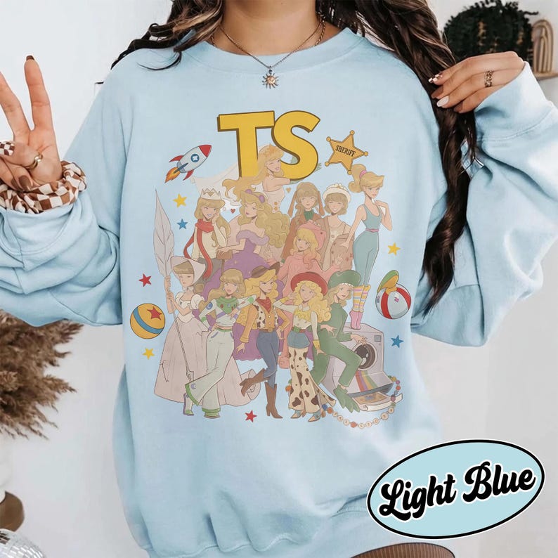 May include: A light blue crewneck sweatshirt featuring a large graphic print of multiple animated characters in various outfits. The text TS appears at the top in yellow, accompanied by a rocket, a sheriff star, and colorful stars.