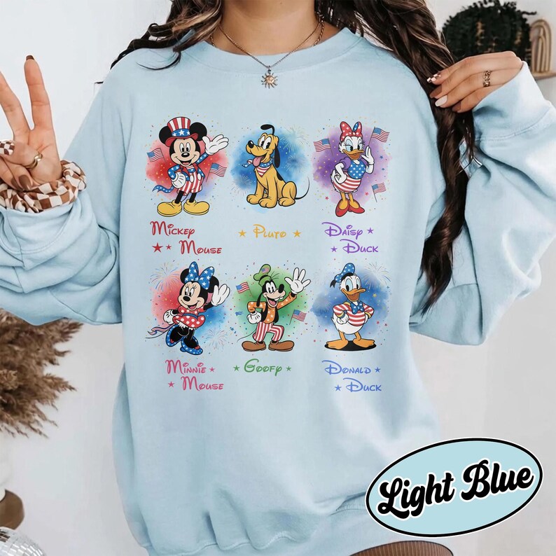 May include: Pale blue sweatshirt with six Disney characters: Mickey Mouse, Pluto, Daisy Duck, Minnie Mouse, Goofy, and Donald Duck. Each character is shown in patriotic outfits with fireworks. The text "Light Blue" is at the bottom.