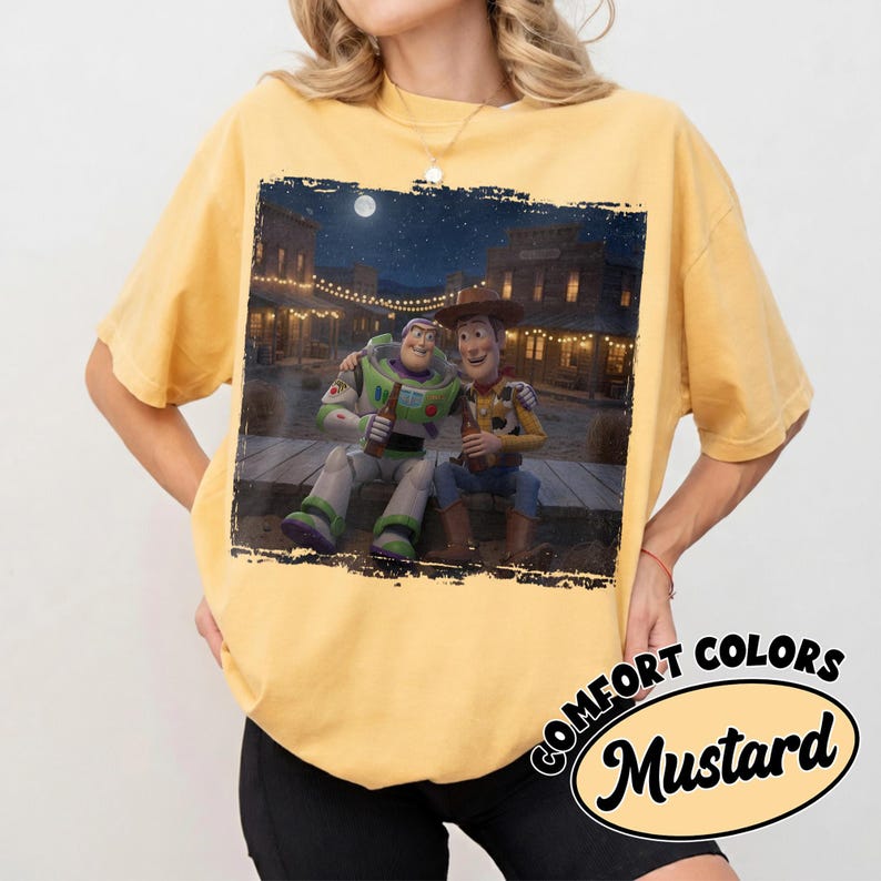 May include: A mustard yellow short-sleeve t-shirt featuring a rectangular graphic print of Buzz Lightyear and Woody from Toy Story sitting together on a wooden porch at night in a western town setting, each holding a brown glass bottle.
