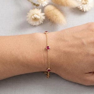May include: A delicate gold chain bracelet featuring three round, vibrant red gemstones. The bracelet is displayed on a wrist, showcasing the jewelry's elegant design and color contrast. The gemstones are set in a bezel setting.