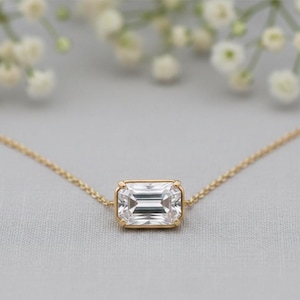 May include: A gold necklace featuring a rectangular, clear gemstone pendant. The pendant is set in a gold bezel and hangs from a delicate gold chain. The background is a soft gray with blurred white flowers.