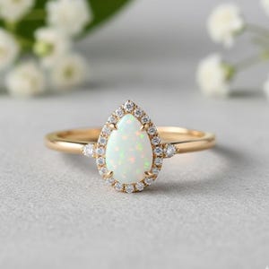 May include: A gold ring featuring a pear-shaped opal gemstone surrounded by a halo of small diamonds. Additional diamonds are set on either side of the opal. The ring is set against a soft, blurred background.