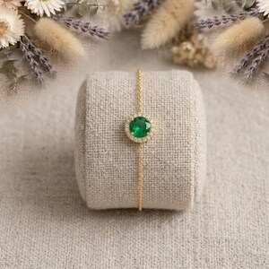 May include: A gold bracelet featuring a round, emerald-green gemstone surrounded by a halo of smaller, clear stones. The bracelet is displayed on a beige fabric roll, with dried flowers in the background. The jewelry is delicate and elegant.