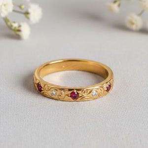 Könnte beinhalten: Ein goldener Bandring mit einem gravierten floralen Muster. Der Ring hat abwechselnd rote und klare Edelsteine. Der Ring ist vor einem weichen, neutralen Hintergrund mit weißen Blumen in der oberen linken Ecke platziert.