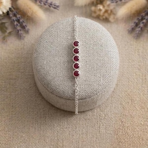 Pode incluir: Uma pulseira de prata com cinco pedras preciosas redondas, vermelho escuro. A pulseira é exibida em uma superfície cinza texturizada, com um fundo macio e neutro. As pedras são espaçadas uniformemente na delicada corrente.