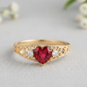 May include: A gold ring featuring a heart-shaped, faceted red gemstone as the centerpiece. The ring is adorned with two smaller, round, clear gemstones on either side of the heart. The band has an intricate, openwork design.