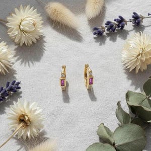 Puede incluir: Un par de pendientes de aro dorados con piedras rosas rectangulares y pequeñas piedras transparentes. Los pendientes se exhiben sobre una superficie gris claro, rodeados de flores secas y vegetación.