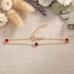 May include: A delicate gold bracelet featuring three round, bezel-set red gemstones. The bracelet has a fine chain and an adjustable clasp. The jewelry is displayed on a neutral linen surface, with dried flowers in the background.