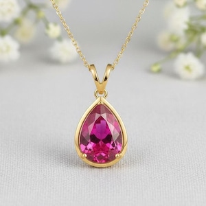 May include: A gold pendant necklace featuring a teardrop-shaped, faceted, pink gemstone. The gemstone is set in a gold bezel and hangs from a delicate gold chain. The background is a soft, neutral color with blurred white flowers.