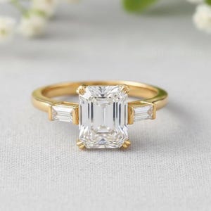 May include: A gold ring with a large, rectangular, clear gemstone as the centerpiece. Two smaller, rectangular gemstones flank the main stone. The ring is set on a light gray surface.