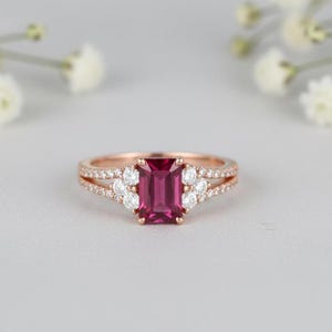 Può includere: Anello in oro rosa con una gemma rettangolare rosa intenso come elemento centrale. L'anello è impreziosito da gemme più piccole, rotonde e trasparenti e da un design a fascia divisa. L'anello è posto su uno sfondo morbido e neutro.