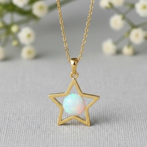 Könnte beinhalten: Eine goldfarbene Halskette mit einem sternförmigen Anhänger. Der Anhänger hat eine Sternkontur und einen runden, opalisierenden Stein. Die Halskette wird auf einer hellgrauen Oberfläche mit weißen Blumen im Hintergrund gezeigt. Die Kette ist ca. 45 cm lang.