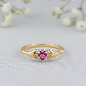 Könnte beinhalten: Ein goldfarbener Ring mit einem herzförmigen, pinkfarbenen Edelstein in der Mitte, umgeben von kleinen, klaren Steinen. Das Design des Rings umfasst stilisierte Hände, die das Herz halten, mit aufwendigen Details am Band.