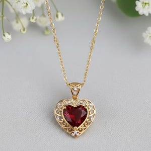 May include: A gold necklace with a heart-shaped pendant. The pendant has a large, deep red gemstone set in a filigree heart design, with small, clear gemstones. The necklace chain is delicate and gold-coloured.