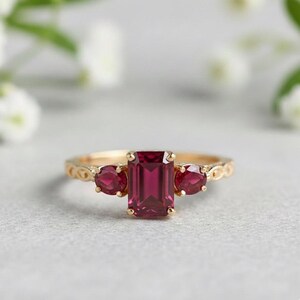 Può includere: Un anello d'oro con una pietra centrale rettangolare rosso intenso, affiancata da due pietre rosse ovali più piccole. L'anello ha una fascia decorativa e strutturata.