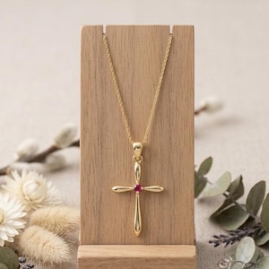 May include: A gold-colored pendant necklace featuring a cross design with a small, red gemstone at the center. The cross is suspended from a delicate chain. The necklace is displayed on a wooden stand against a soft, neutral background.