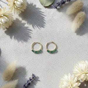 May include: A pair of gold hoop earrings, each adorned with three emerald-green rectangular stones. The earrings are displayed on a textured white surface, surrounded by dried flowers and foliage, creating a natural aesthetic.