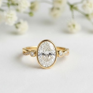 May include: A gold engagement ring with a large oval-cut diamond center stone. The band features smaller diamonds and a delicate design. The ring is set against a soft white background.