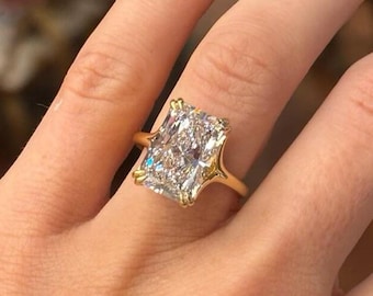 May include: A radiant-cut diamond ring with a gold band. The large, rectangular diamond is set in a gold prong setting. The ring is on a person's hand with manicured nails.