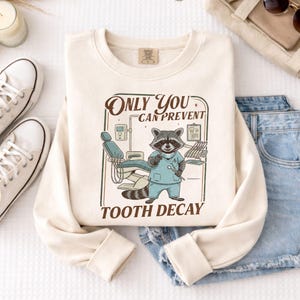 Op de afbeelding: Cr&egrave;mekleurige sweatshirt met een afbeelding van een wasbeer als tandarts, met een tandheelkundig instrument. De tekst luidt "Only You Can Prevent Tooth Decay". Het ontwerp omvat een tandartsstoel en gereedschap. De sweatshirt is op een wit oppervlak gevouwen.