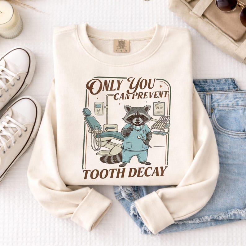 Op de afbeelding: Cr&egrave;mekleurige sweatshirt met een afbeelding van een wasbeer als tandarts, met een tandheelkundig instrument. De tekst luidt "Only You Can Prevent Tooth Decay". Het ontwerp omvat een tandartsstoel en gereedschap. De sweatshirt is op een wit oppervlak gevouwen.