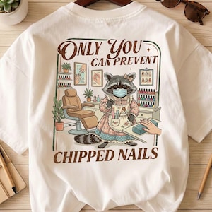 Op de afbeelding: Wit T-shirt met een cartoon wasbeer in een nagelsalon. De wasbeer draagt een gezichtsmasker en schort, met gereedschap. Het shirt heeft de tekst "Only You Can Prevent Chipped Nails" in een bruin, vintage lettertype.