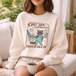 Op de afbeelding: Cr&egrave;mekleurige sweatshirt met een cartoon wasbeer in een tandartsuniform, staand voor een tandartsstoel. De tekst luidt "Only You Can Prevent Tooth Decay". Het ontwerp staat in een rechthoekige lijst.
